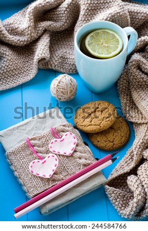 A cup of tea in sweater with lemon, old notebook with hearts of felt, beige knitted blanket and spokes lie on blue background