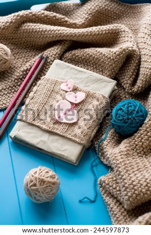 One old notebook in knitted cover with felt hearts lie next to the coil bright filaments and blanket knitted on blue background