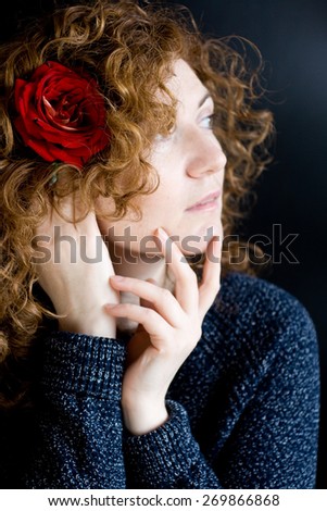 Portrait of beautiful young redhead with curly red rose in her hair on a dark background