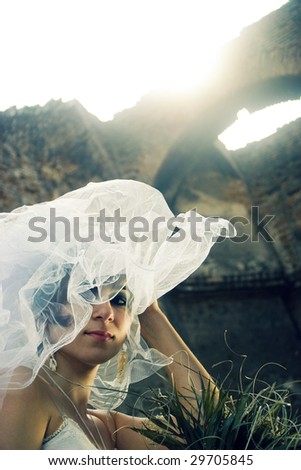 Bride portrait with her bouquet in her hands and white veil in the wind and sun, the ruins of a church in the background
