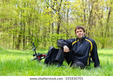 Happy man cyclist with bike and backpack sitting on green grass against the background of nature in spring on a sunny day