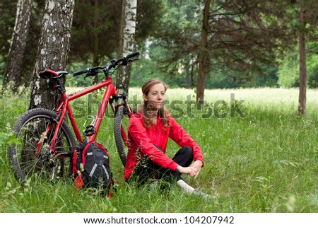 Woman cyclist with bike and backpack in a red tracksuit relaxing among of green nature in spring in the park