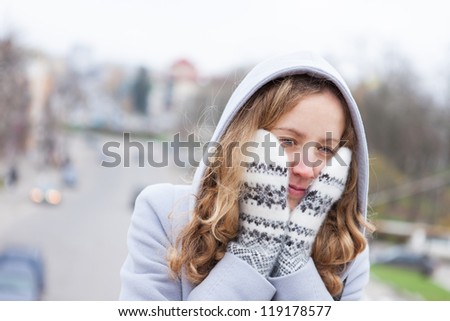 Frozen young woman heated by wool mittens and a from hood against the blurred background city. Outdoor