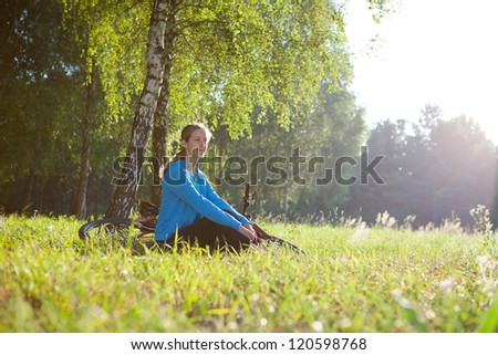 Woman cyclist enjoying relaxation in spring sunny park sitting in the fresh green grass near a birch trunk