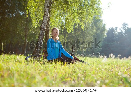 Young woman cyclist enjoying relaxation in spring sunny park sitting near a birch trunk
