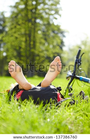 Happy cyclist enjoying relaxation lying barefoot in green grass outdoors in summer park