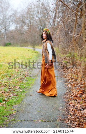 Young girl with flowers wreath in fur coat goes along the asphalt path in a autumn park