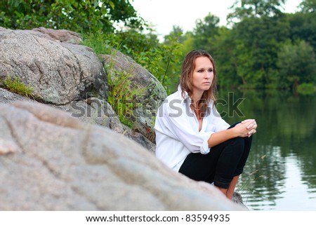 A lonely young woman sitting on rocks on the river arms folded on her lap and staring into the distance