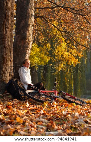 Relaxes woman cyclist with bike sits among fallen leaves in autumn park illuminated by the rays sun and enjoy recreation