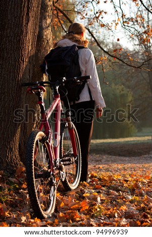 Woman cyclist with bike and backpack in autumn nature on the morning, lit morning sun
