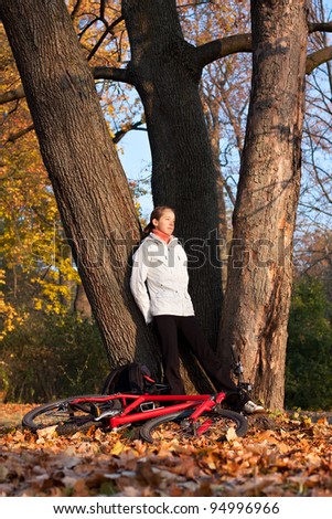 Relaxes woman cyclist with bike standing among old trees in autumn park illuminated by the rays sun and enjoy recreation