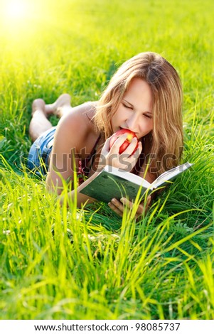 Happy beautiful woman with an apple in hand lying on the green grass and reading a book a sunny day against a background of green nature