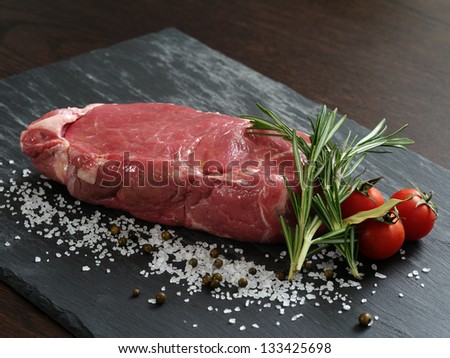 Photo of a raw thick sirloin steak with rosemary, cherry tomatoes, salt and peppercorns on a piece of black slate.