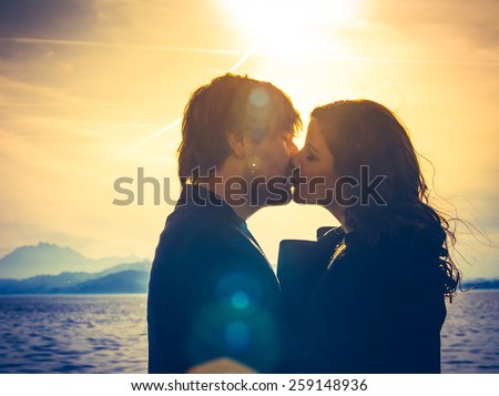 Photo of young adults by the water hugging and kissing in the late afternoon as the sun lowers in the sky. Heavily filtered for more romantic effect. Real lens flare visible.
