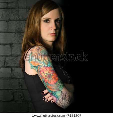 A young female with full arm tattoo leaning up against a black brick wall.