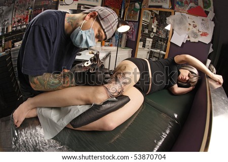A tattoo artist applying his craft onto the leg of a female. Property release supplied includes tattooists\' studio hanging artwork and his tattoo work. Shot with fisheye lens.