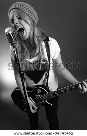 Black and white photo of a blond female screaming into an old microphone and playing electric guitar.  High contrast with film grain added.