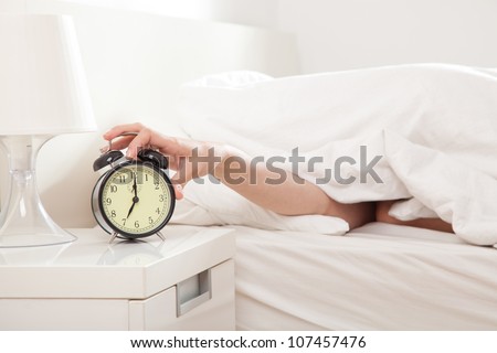 Hand under blanket reaching out for alarm clock, shallow depth of field focus on foreground