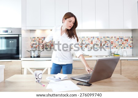 Beautiful young woman working on laptop and cooking at the same time