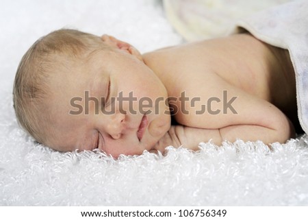 Closeup image of a six-day old newborn sleeping peacefully on a fluffy white blanket and covered, waste down, with a pastel blanket.