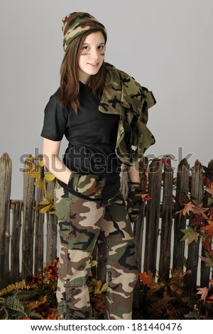 A beautiful young teen by a rustic old fence.  She\'s wearing camo pants, hat, and gloves and carries her jacket over her shoulder.  On a gray background.