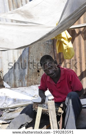 SAINT MARC, HAITI - FEBRUARY 22, 2013:  A Haitian craftsman practicing his trade by his home.  He\'s sitting in the shade that he created by hanging cloths.