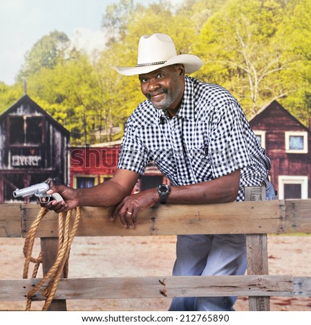 A mature African American cowboy happily looking at the viewer while leaning against a rustic rail fence and holding a gun.