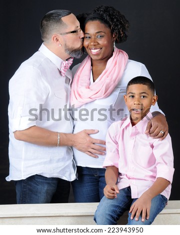 Portrait of a mixed race family.  Profile of the husband kissing his pregnant wife with his hand on her expanding belly while their elementary son sits with the pair.  On a black background.