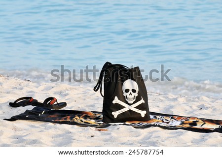 A black beach back with a smiling skull and crossbones with a towel and flip flops  on a sandy beach.