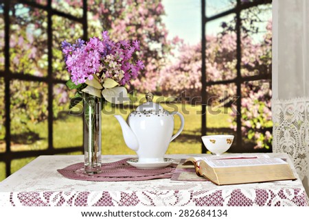 An opened Bible on a table set with a teapot, tea cup and bouquet of violate lilacs, all set before a window opened to a sunny field of lilacs bushes.  Shallow depth of field with focus on the Bible.