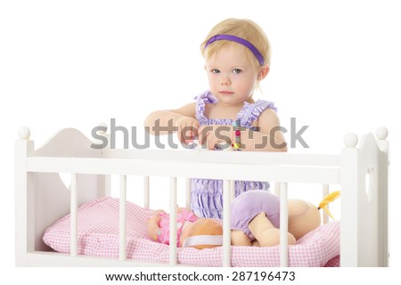 An adorable 2-year-old holding a baby bottle in her hands while standing by her doll crib containing two dolls.  On a white background.