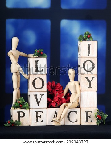 Love, Joy and Peace spelled out with rustic alphabet blocks in front of a night-time window.  Two mannequins, holly and a small bouquet of poinsettias adorn the blocks.