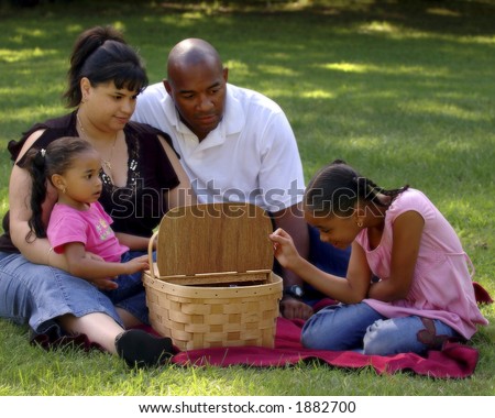 Child peeking into picnic basket with her bi-racial family looking on.