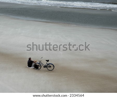 Man resting on an empty beach overlooking the ocean on an overcast day.  A bicycle lays by his side.