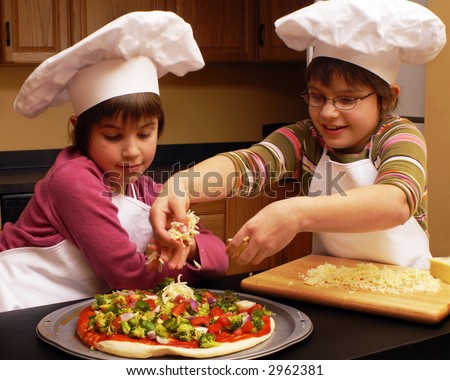 Two sisters in aprons and chef\'s hats making a veggie pizza.