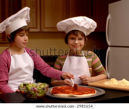Sisters in aprons and chef\'s hats spreading tomato sauce on pizza crust with veggies and cheese close by.