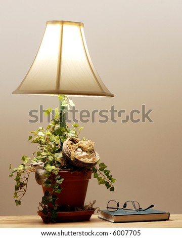 A closed book and eye glasses set under a small flower-pot table lamp decorated with a bird\'s nest,foliage and a bunny.