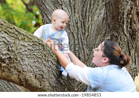 A baby sitting on a big tree limb, laughing at his mother who's securing him.