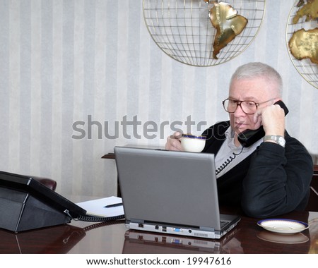 Senior businessman in his office drinking a cup of coffee while talking on the phone and studying his computer.