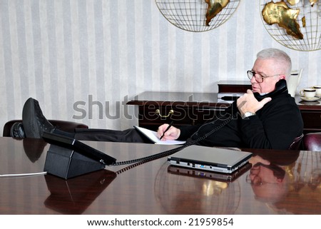 A senior businessman relaxed with his feet up while taking notes from a business phone conversation in the conference room.