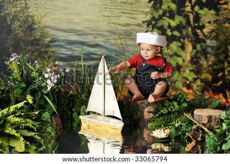 A happy baby boy in a sailor\'s hat playing with a toy sail boat by the water\'s edge.