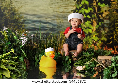 A barefoot baby boy happily playing by the water\'s edge with a rubber duck -- both in sailor\'s hats.