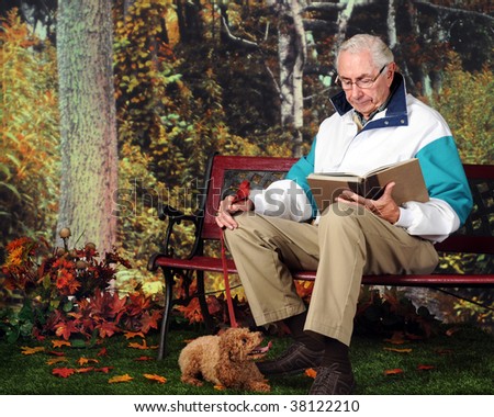 A senior man reading a book on a park bench in the fall; his pet poodle resting at his feet.