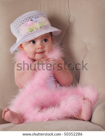An adorable baby girl sitting in a plush chair, wrapped in pink boas, beads and a fancy hat.