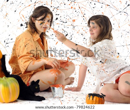 A young teen and her preteen sister making a mess of themselves with black and orange paint.  Pumpkins and a black cat nearby.  Some motion blur on the brush painting the girl\'s face.