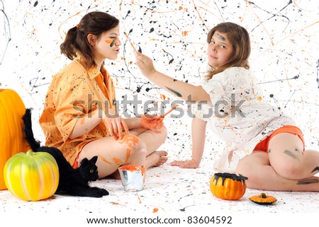 Two sisters deep in black and orange paint, surrounded by pumpkins, a black can and a paint-spattered background.