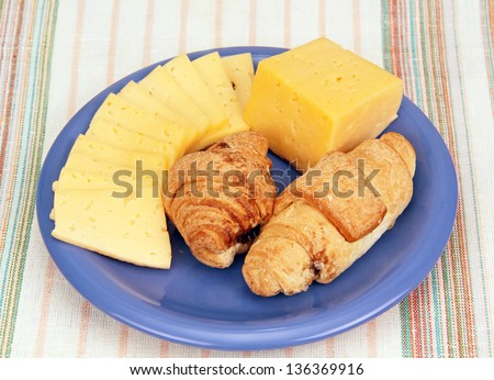 piece of cheese and thin slices of cheese with two croissants on a blue plate.