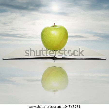 Open book with a green apple above of it in the beautiful sky on the background and its reflection