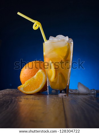 refreshing cocktail with orange in misting glass, orange fruit with slice and ice on a wooden table on dark-blue background