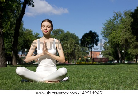 young woman in white closing sitting in yoga pose on green grass in park and meditating,  green trees, building  and blue sky background
